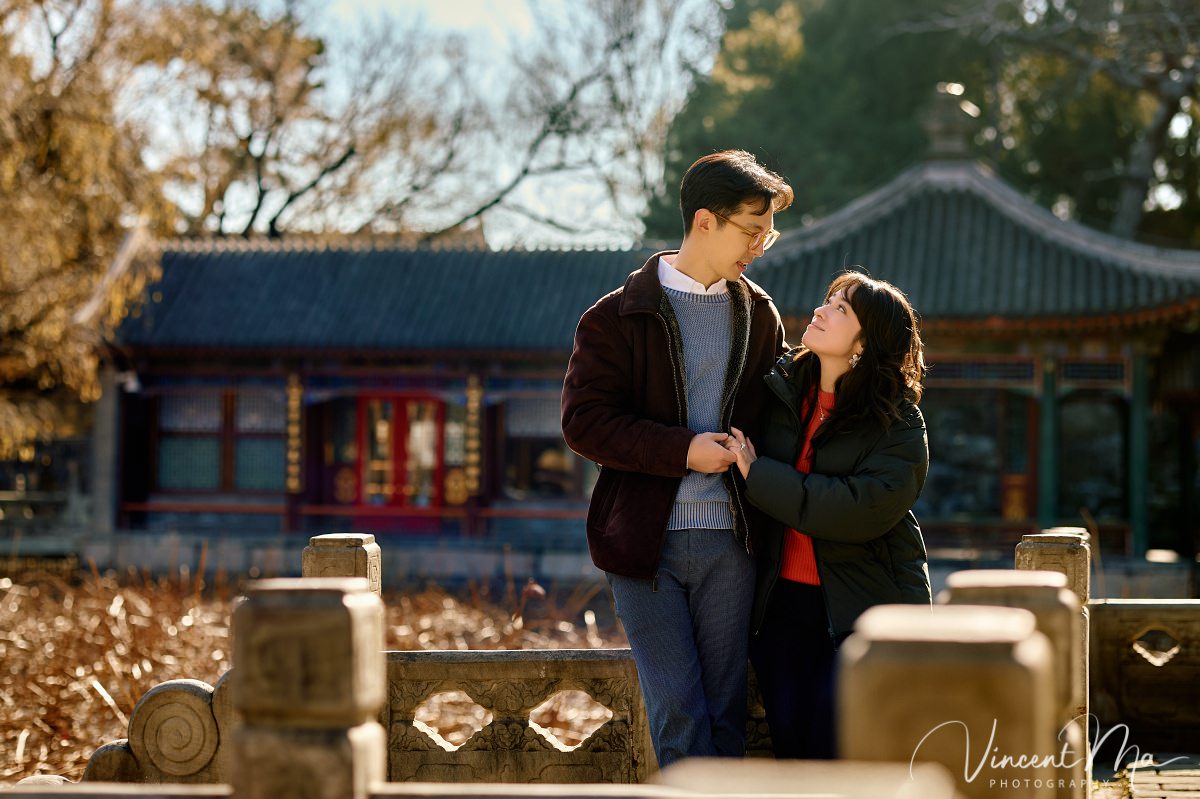 Beijing engagement photography. Couple looking at each other lovingly on a stone bridge at the Summer Palace. Traditional Chinese pavilion and winter reeds in the background.
