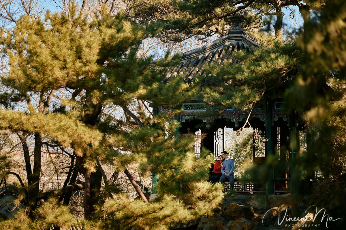 Beijing engagement photography. Couple looking at each other lovingly on a stone bridge at the Summer Palace. Traditional Chinese pavilion and winter reeds in the background.