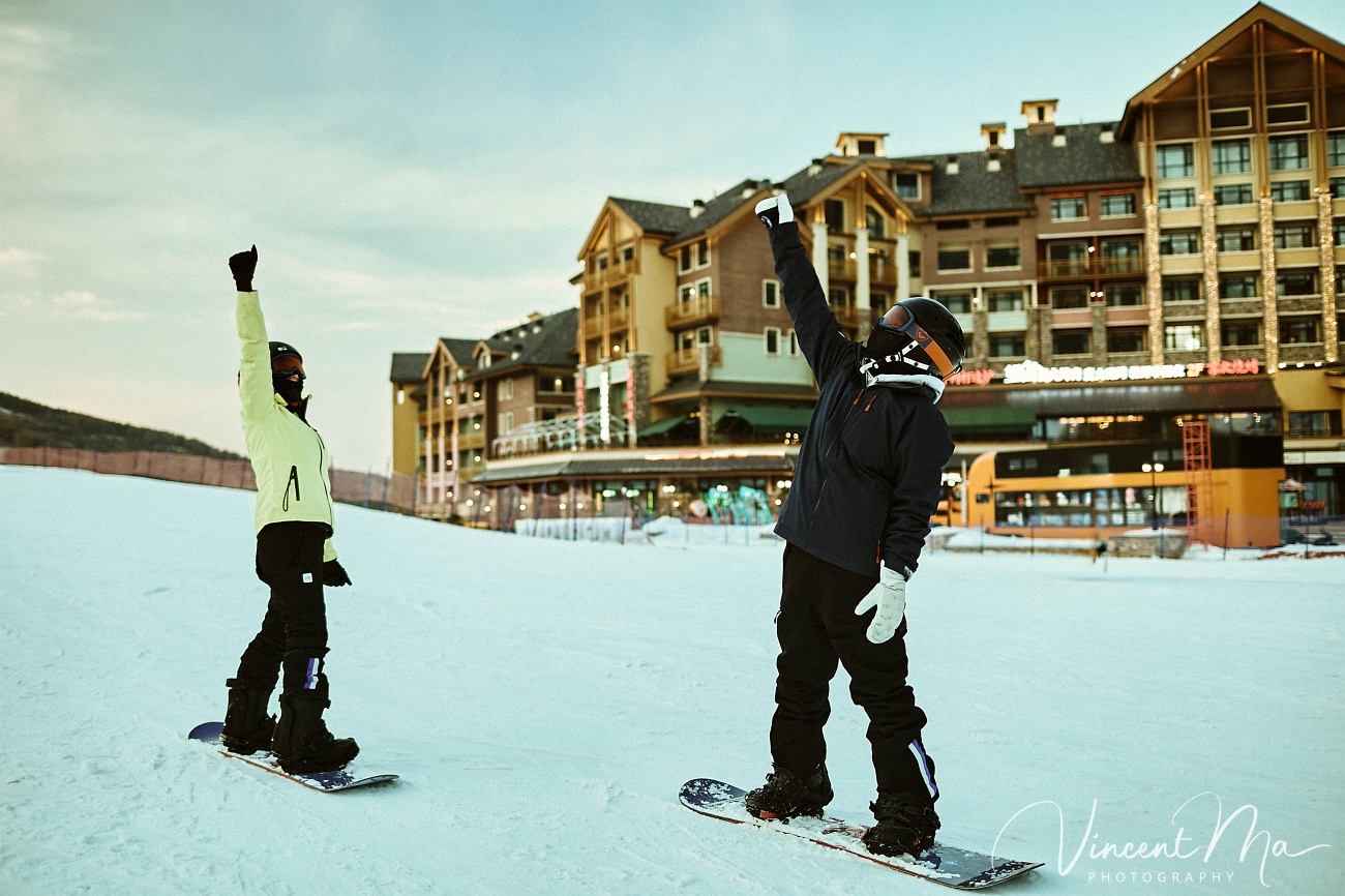 Same-sex couple snowboarding together in powder snow wearing cute turtle and avocado protective gear at a Chongli ski resort.