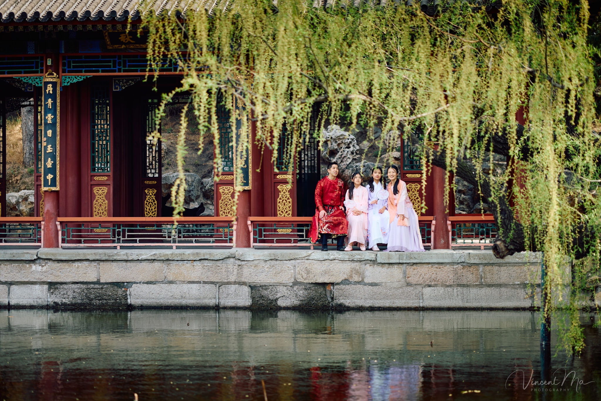 Family of four sitting by the lake under green weeping willows and spring cherry blossoms at the Summer Palace.