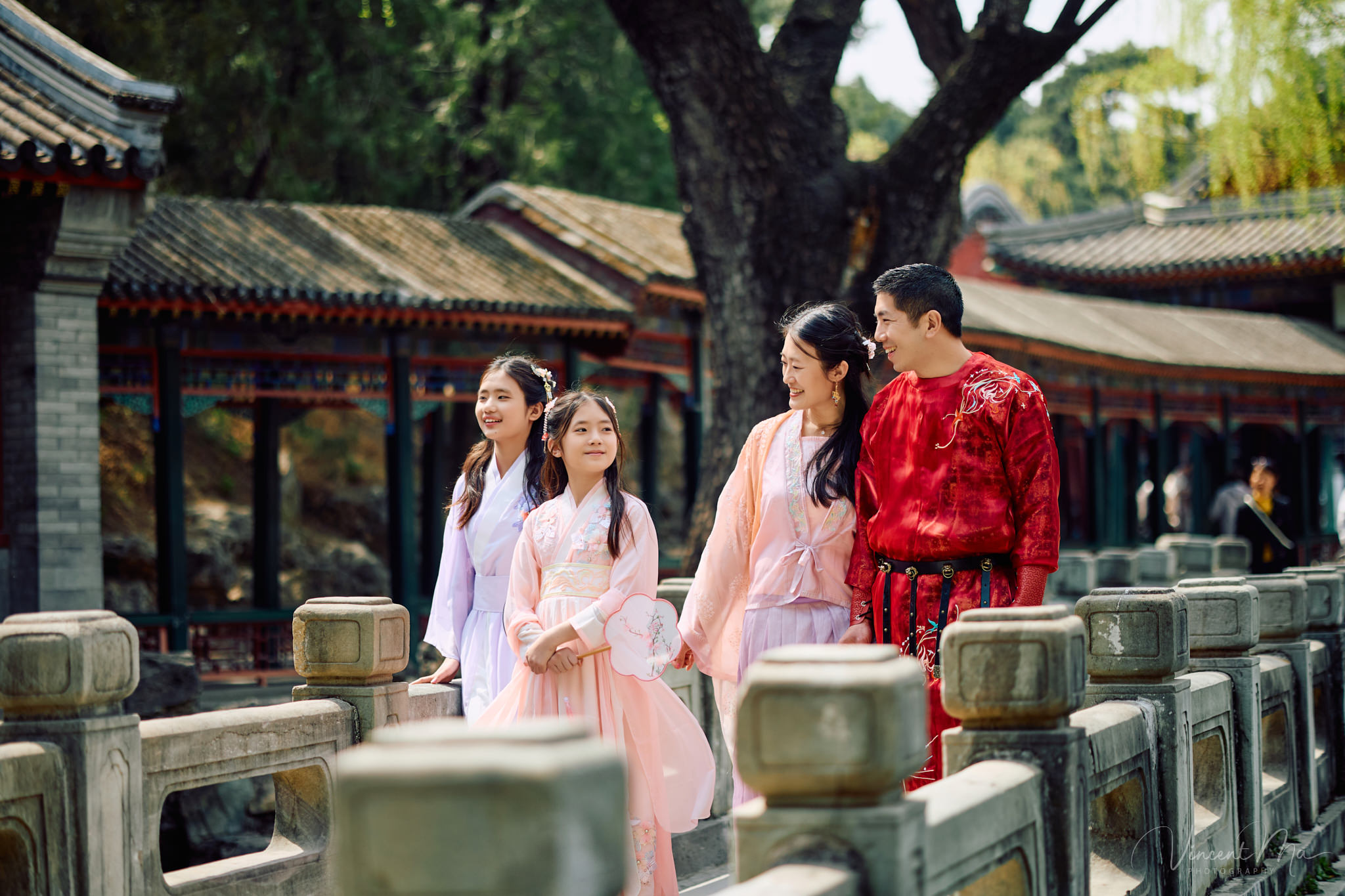 Family of four sitting by the lake under green weeping willows and spring cherry blossoms at the Summer Palace.
