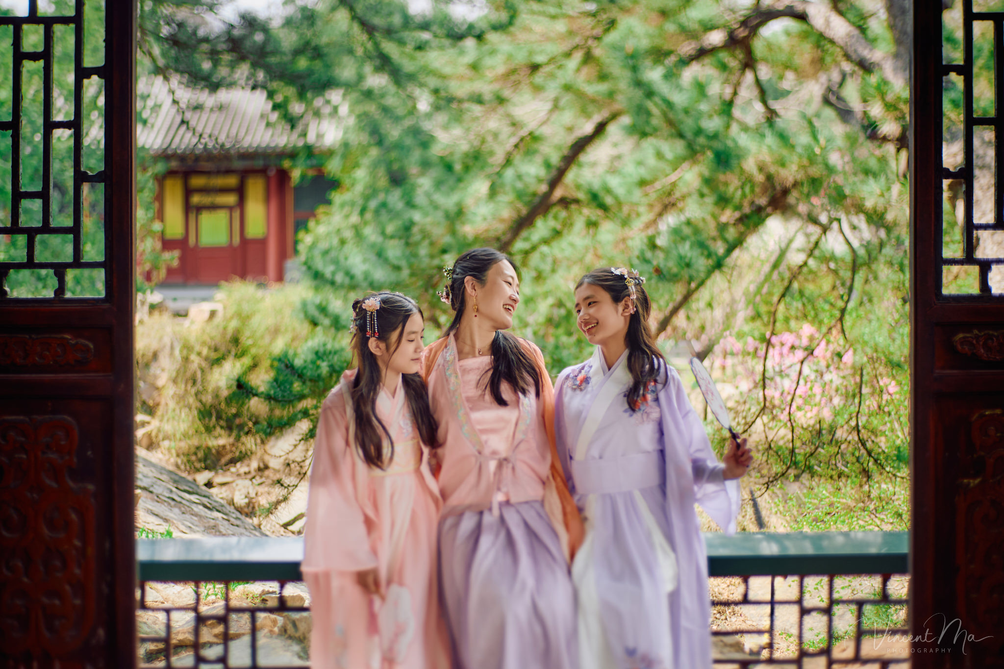 Little girl holding a traditional Chinese fan, Pink peach blossoms in Beijing spring, Family walking on ancient stone path, Vintage wooden window architecture detail