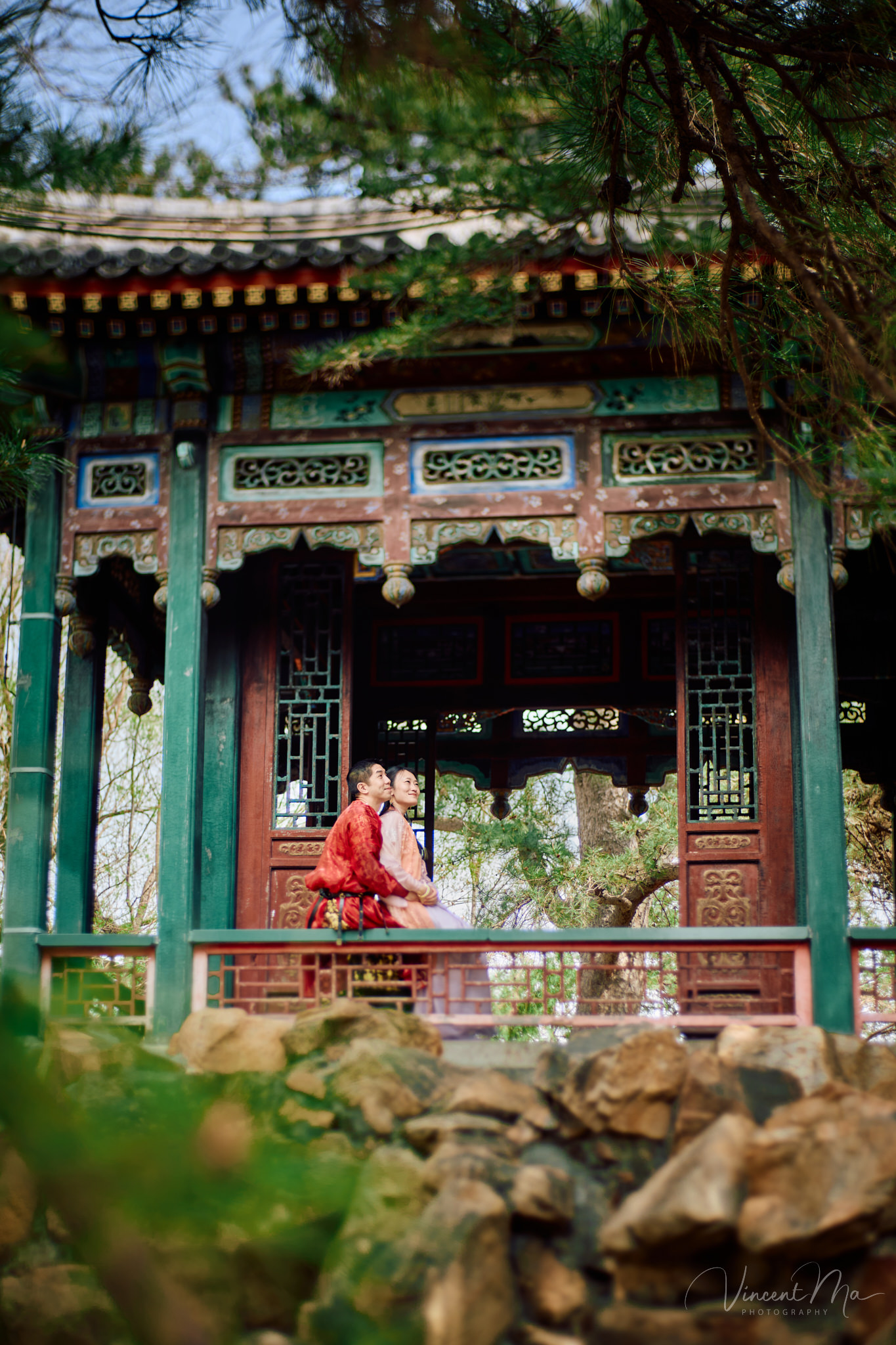American couple in red and pink traditional Chinese clothing framed by delicate apricot blossoms during a Beijing spring photoshoot.