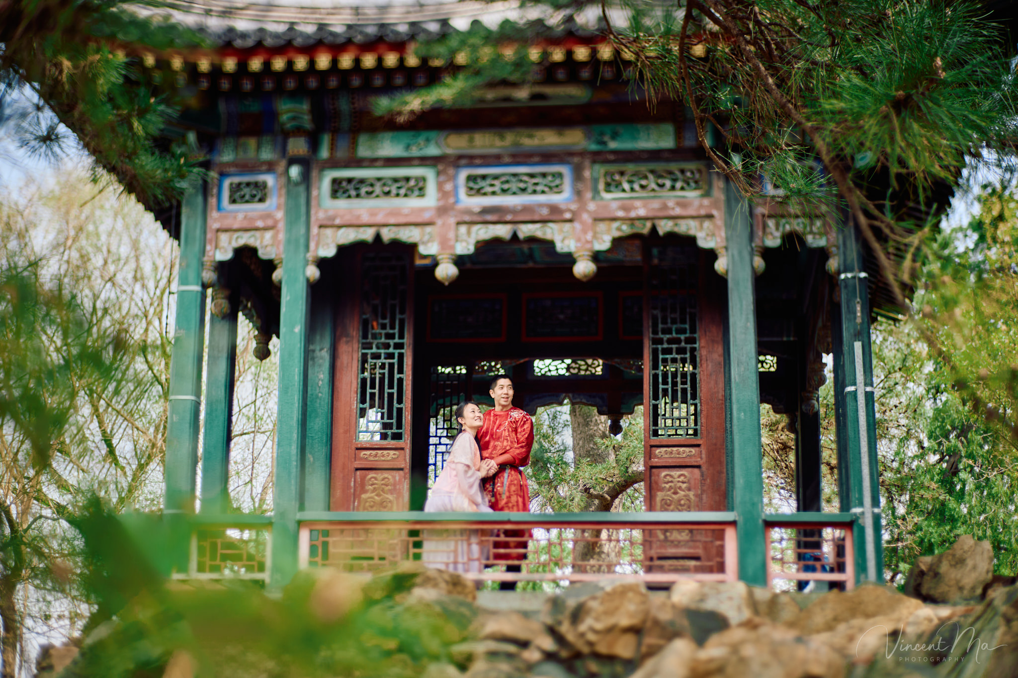 American couple in red and pink traditional Chinese clothing framed by delicate apricot blossoms during a Beijing spring photoshoot.