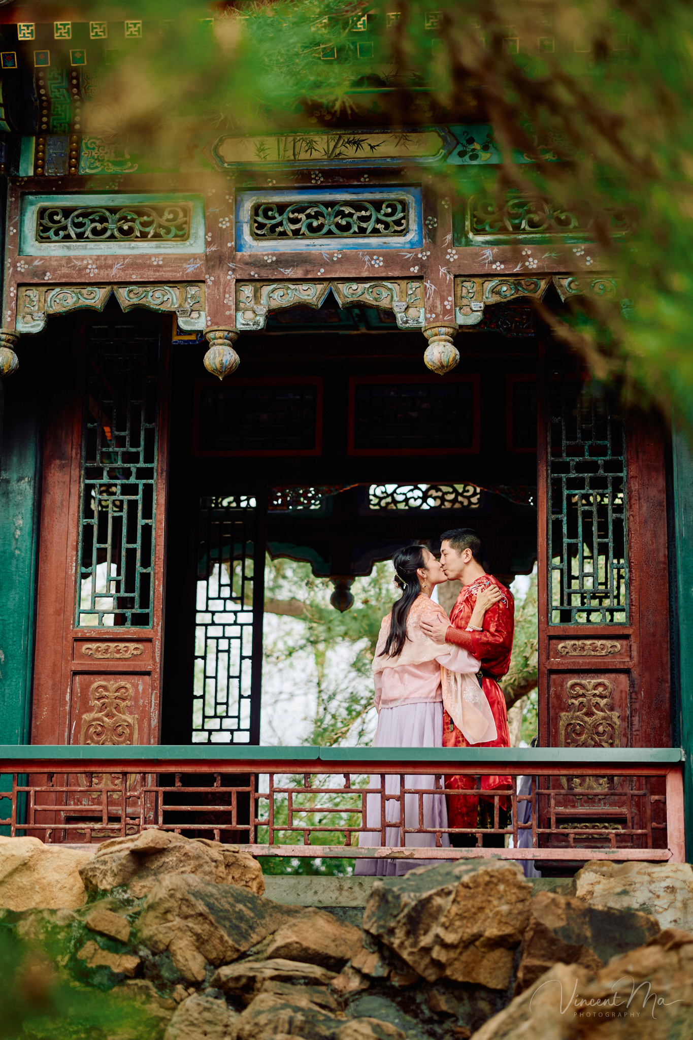 American couple in red and pink traditional Chinese clothing framed by delicate apricot blossoms during a Beijing spring photoshoot in the Summer palace.