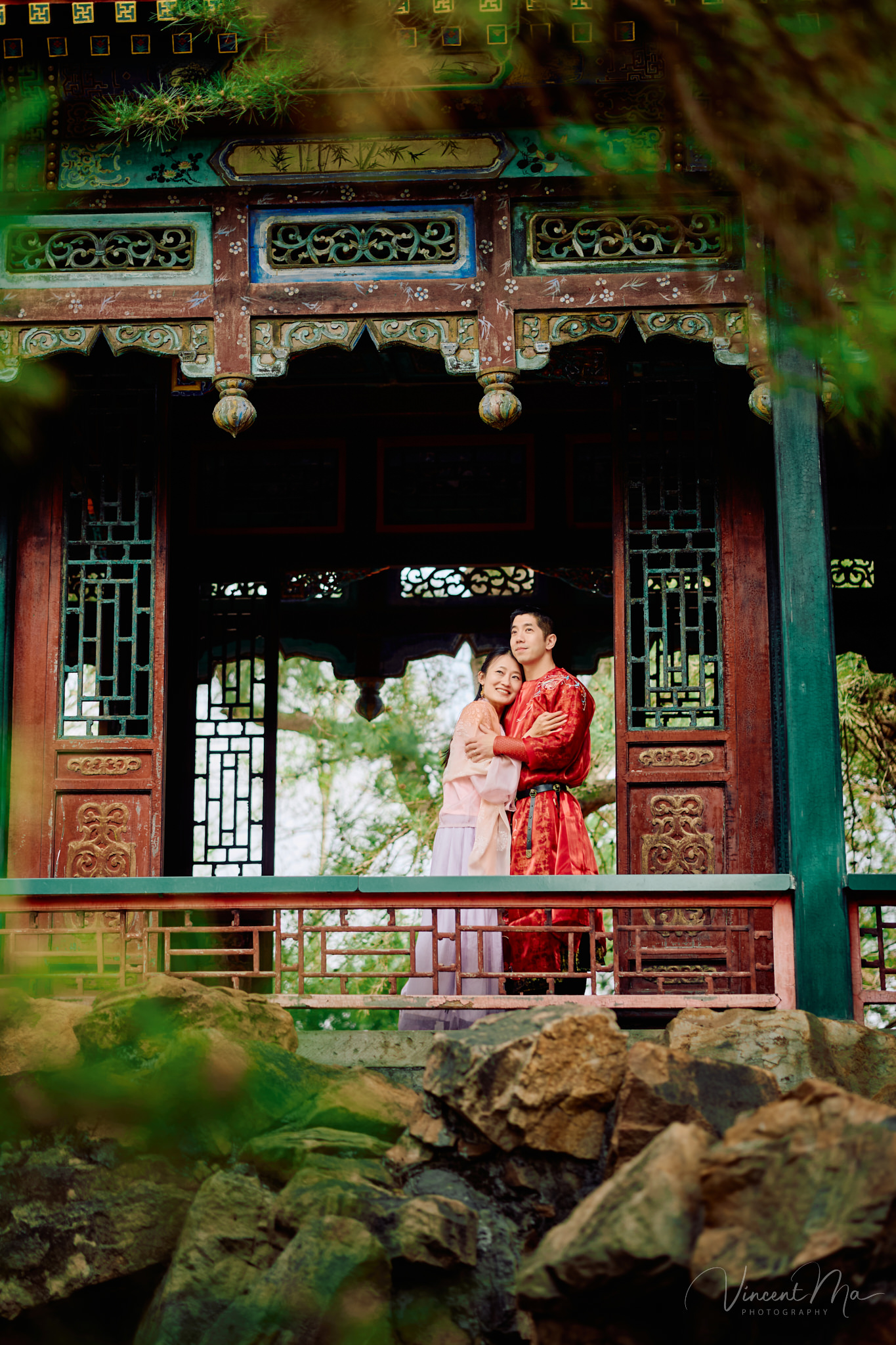American couple in red and pink traditional Chinese clothing framed by delicate apricot blossoms during a Beijing spring photoshoot in the Summer palace.