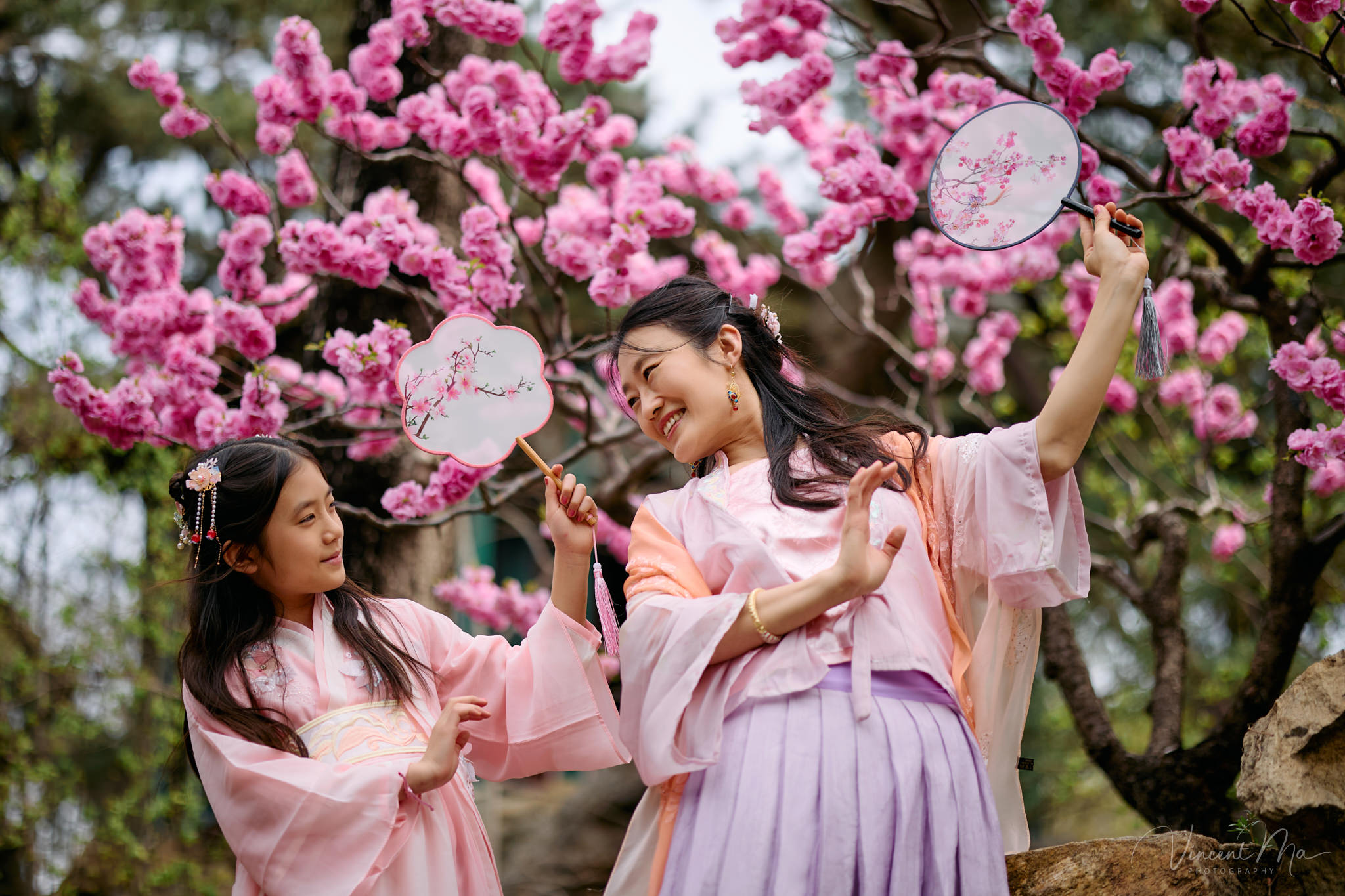 Little girl holding a traditional Chinese fan, Pink peach blossoms in Beijing spring, Family walking on ancient stone path, Vintage wooden window architecture detail