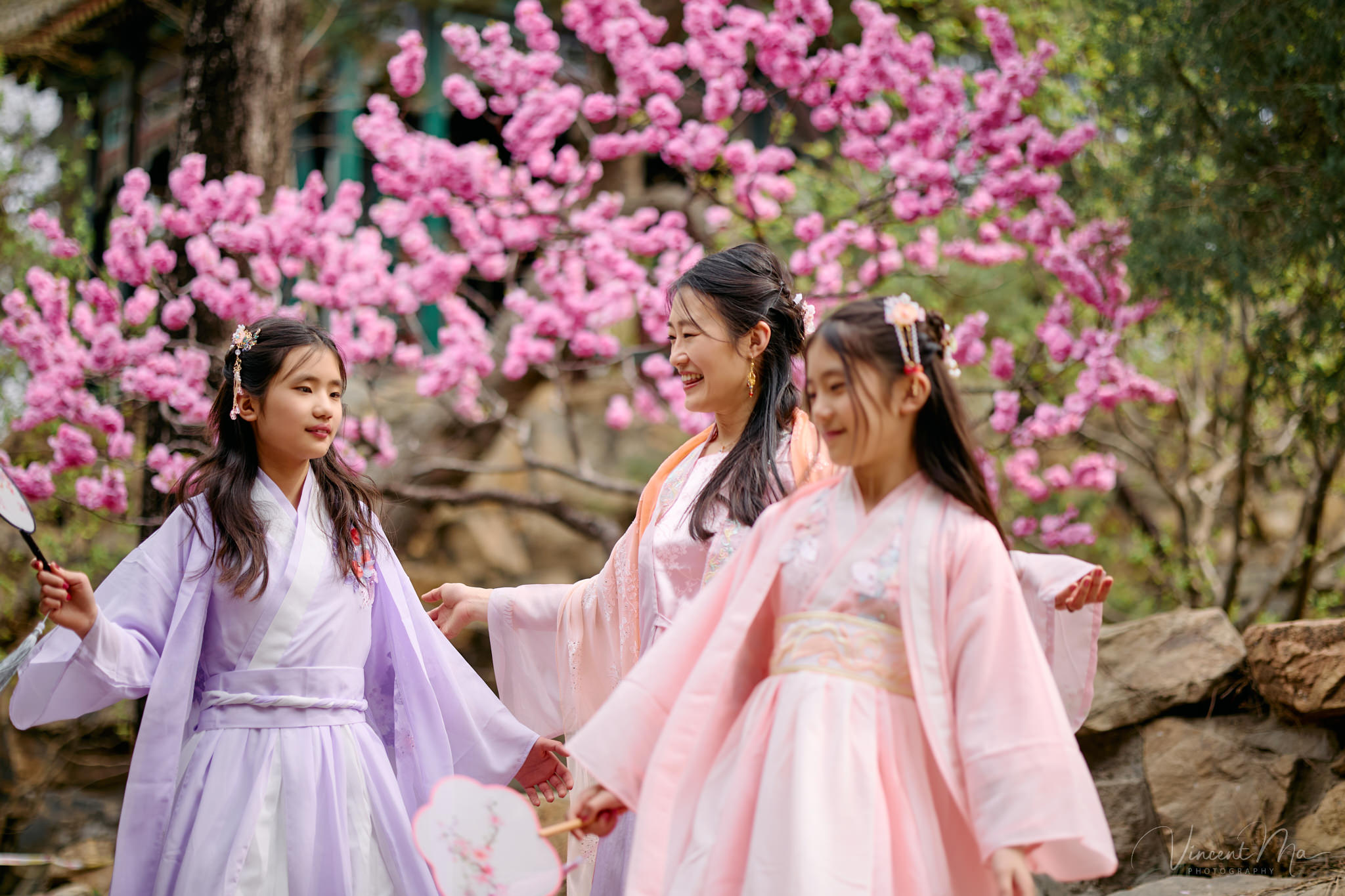 Mother and daughter in pink Hanfu costumes playing with traditional fans under blooming spring peach blossoms at the Summer Palace.