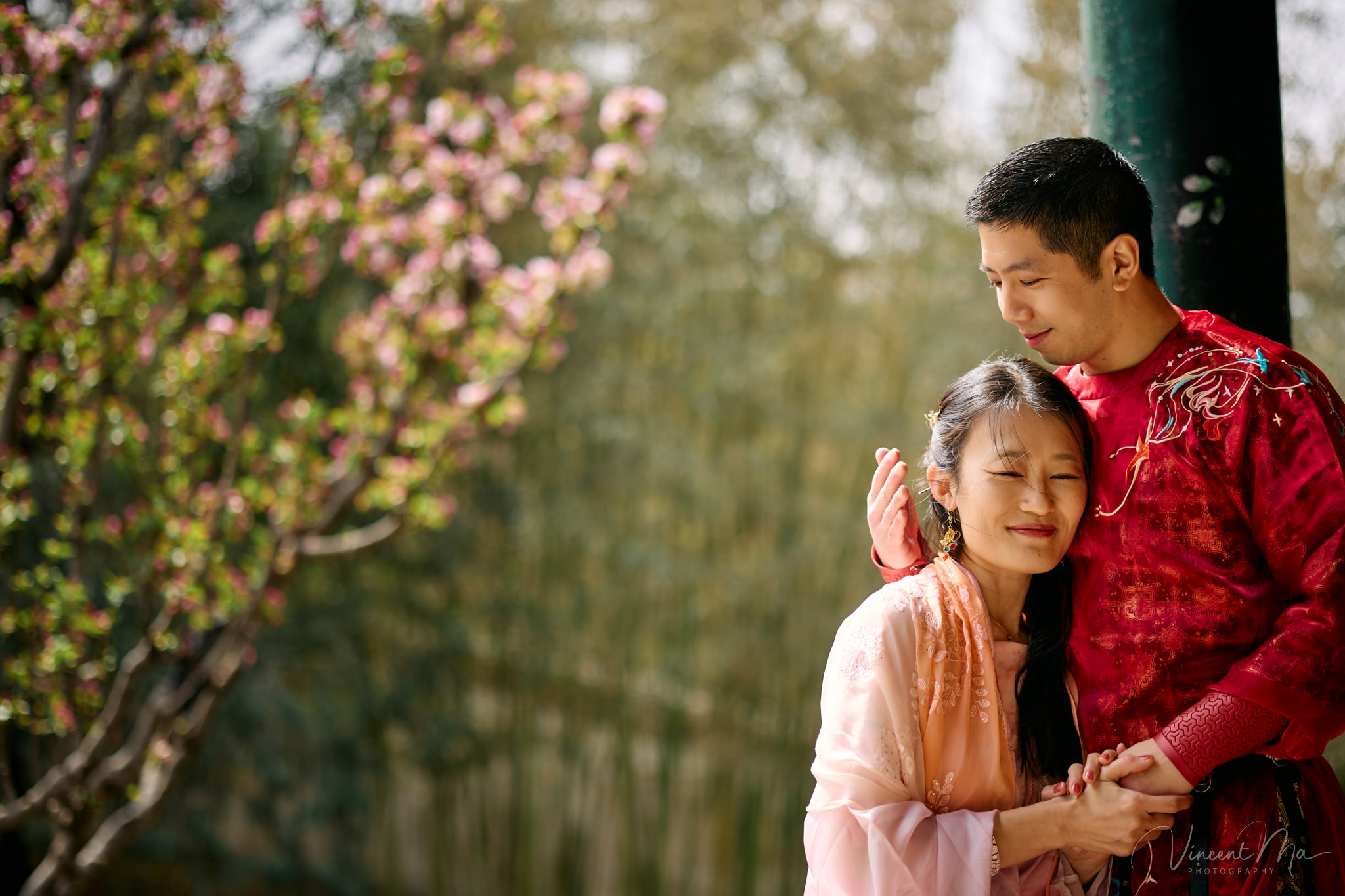 American couple in red and pink traditional Chinese clothing framed by delicate apricot blossoms during a Beijing spring photoshoot in the Summer palace.