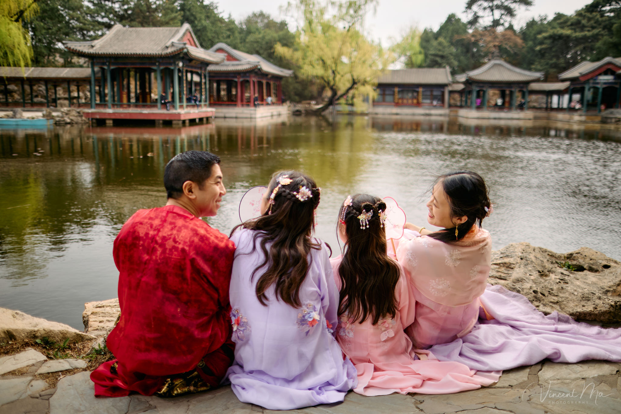 Family of four sitting by the lake under green weeping willows and spring cherry blossoms at the Summer Palace.