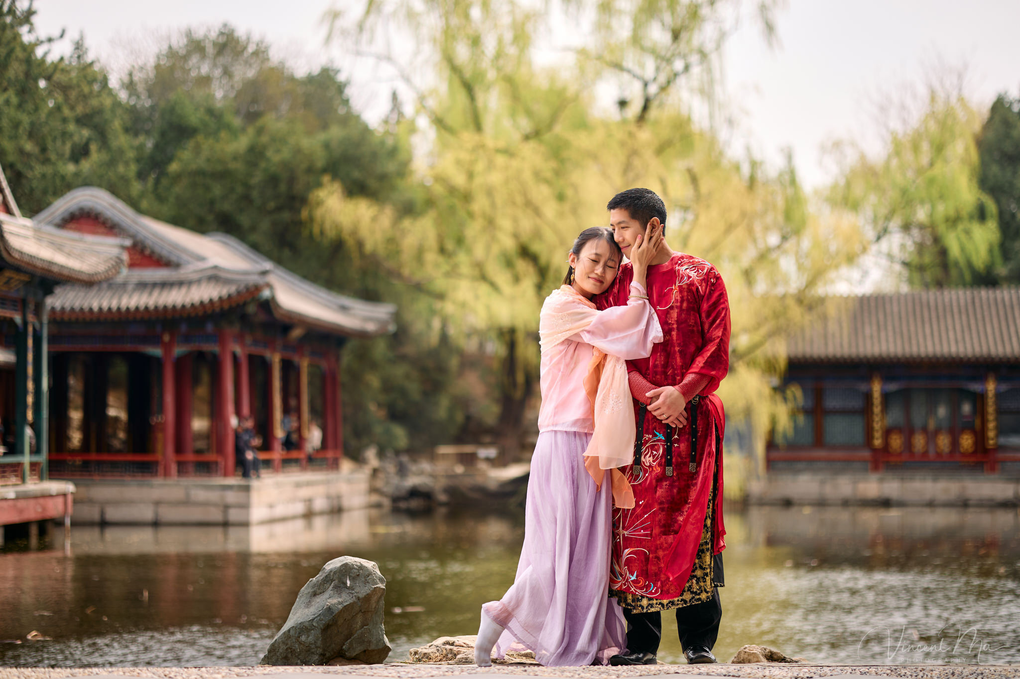 American couple in red and pink traditional Chinese clothing framed by delicate apricot blossoms during a Beijing spring photoshoot in the Summer palace.