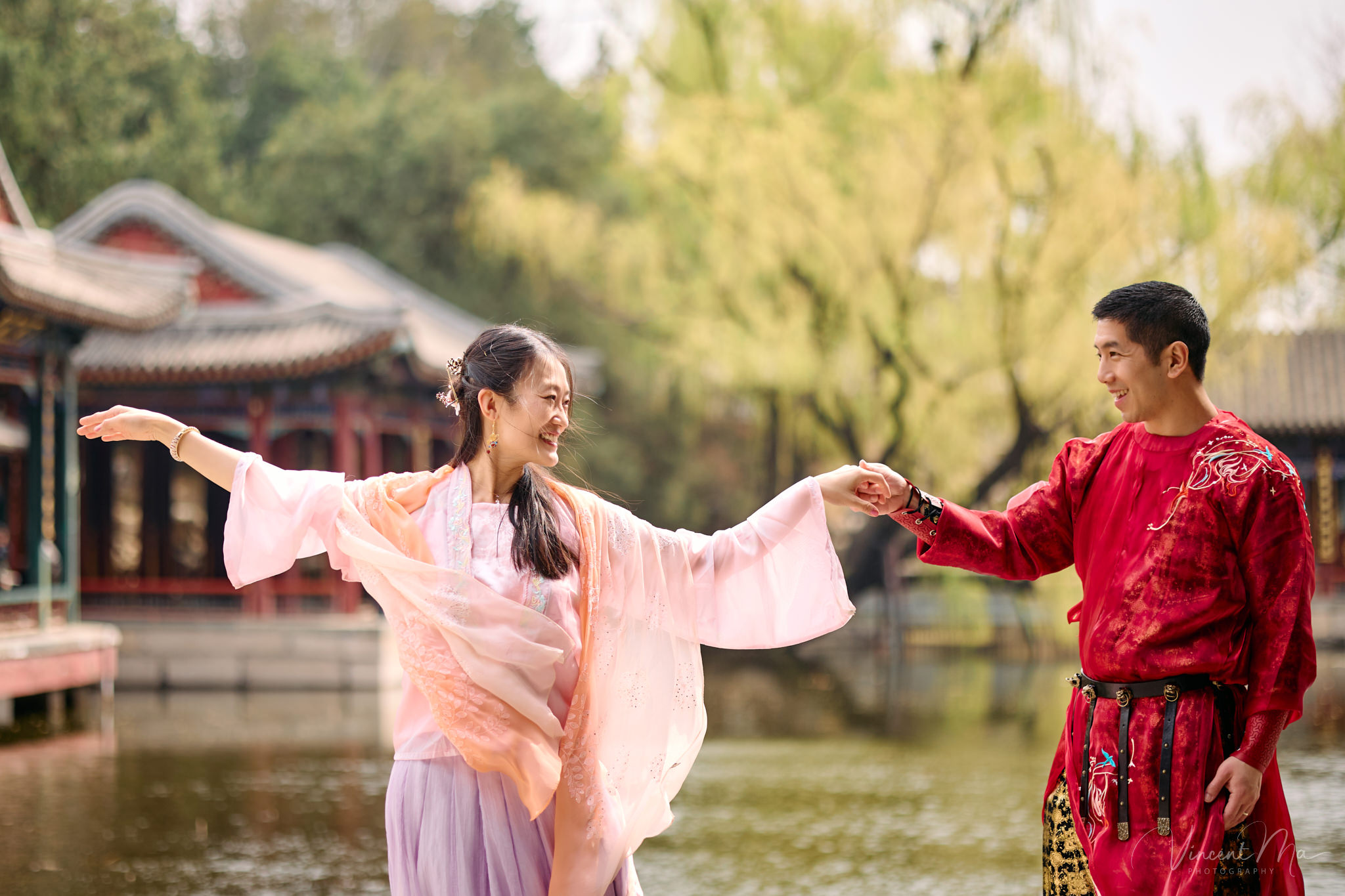 American couple in red and pink traditional Chinese clothing framed by delicate apricot blossoms during a Beijing spring photoshoot in the Summer palace.