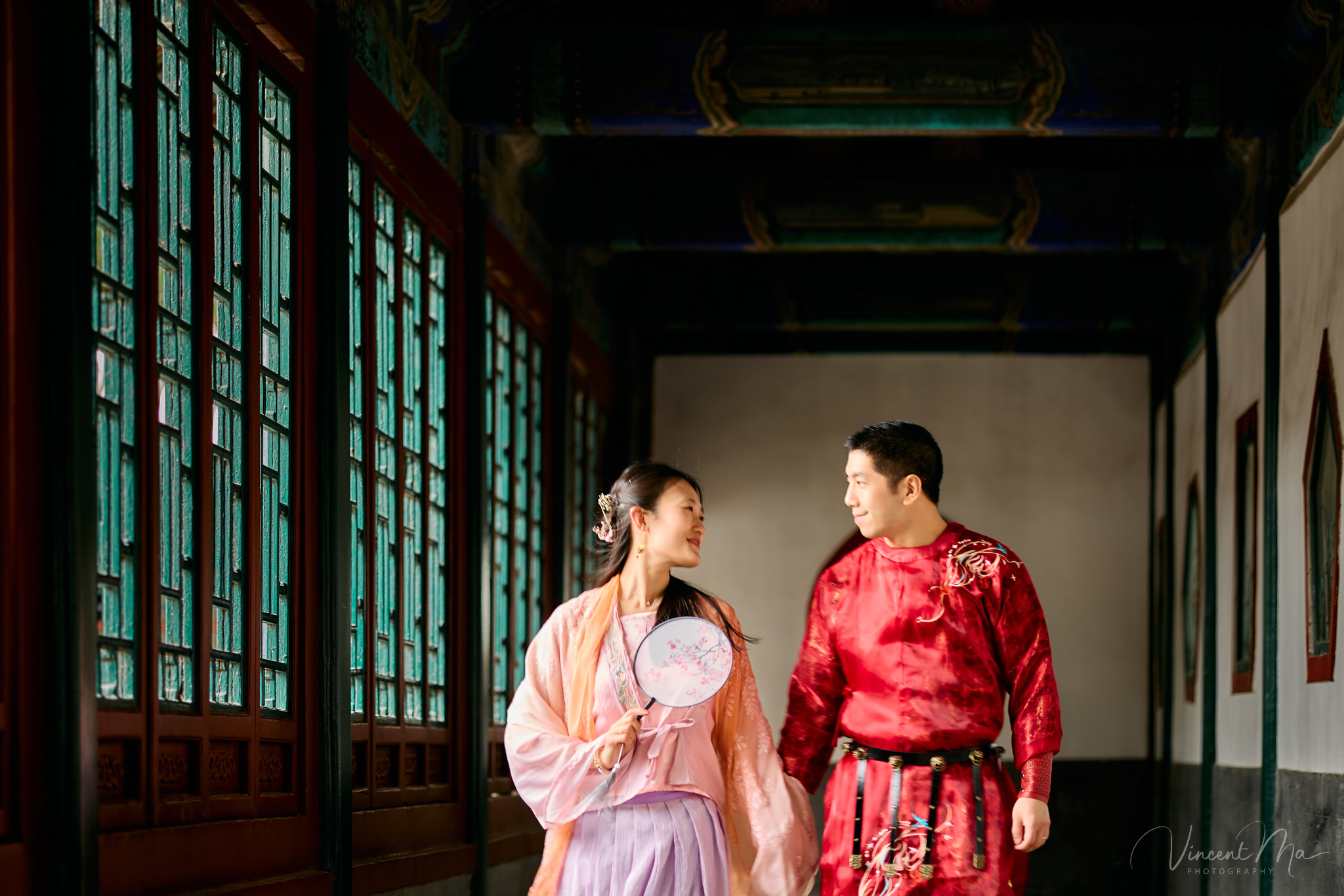 American couple in red and pink traditional Chinese clothing framed by delicate apricot blossoms during a Beijing spring photoshoot in the Summer palace.