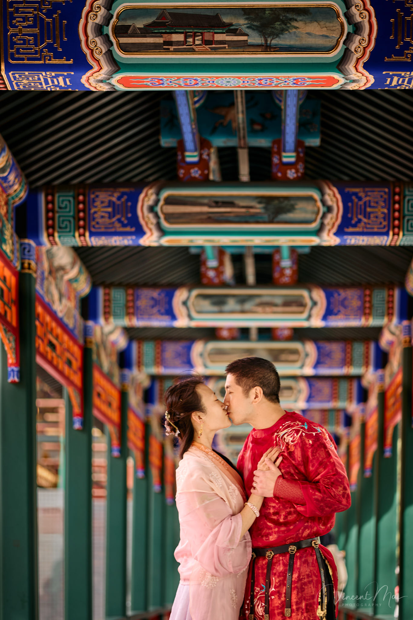 American couple in red and pink traditional Chinese clothing framed by delicate apricot blossoms during a Beijing spring photoshoot in the Summer palace.