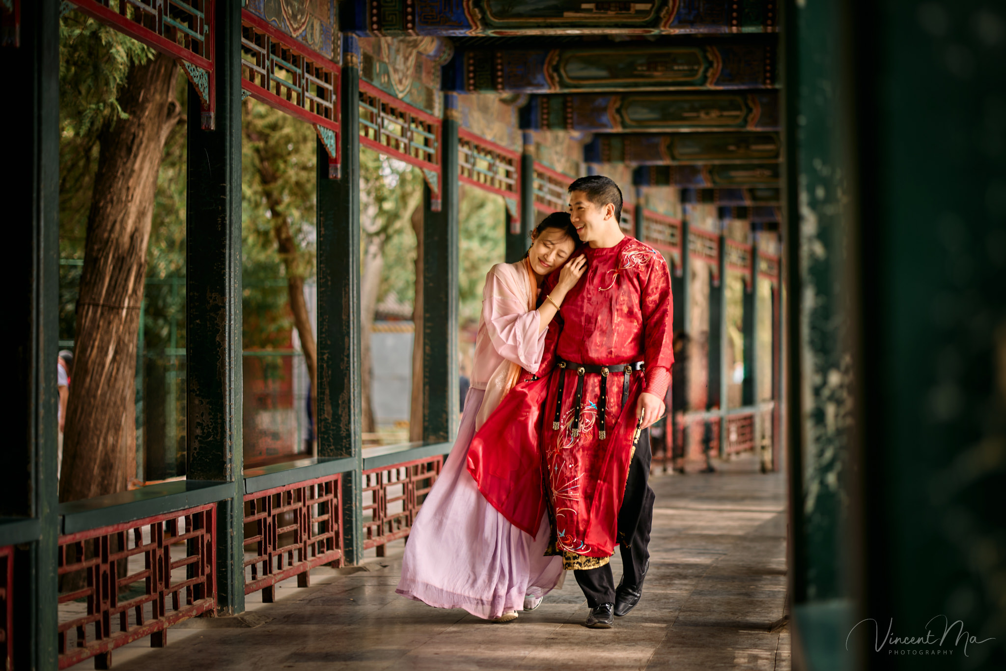 American couple in red and pink traditional Chinese clothing framed by delicate apricot blossoms during a Beijing spring photoshoot in the Summer palace.