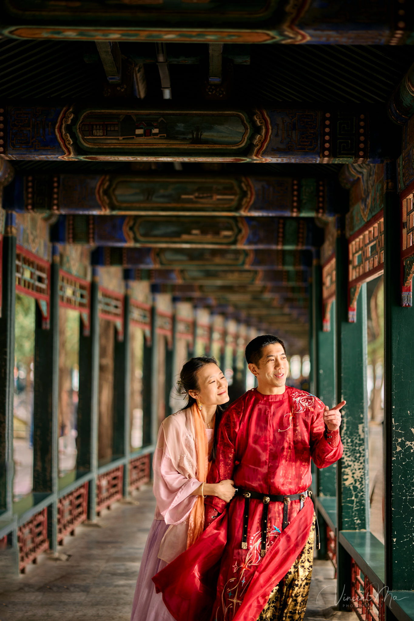American couple in red and pink traditional Chinese clothing framed by delicate apricot blossoms during a Beijing spring photoshoot in the Summer palace.