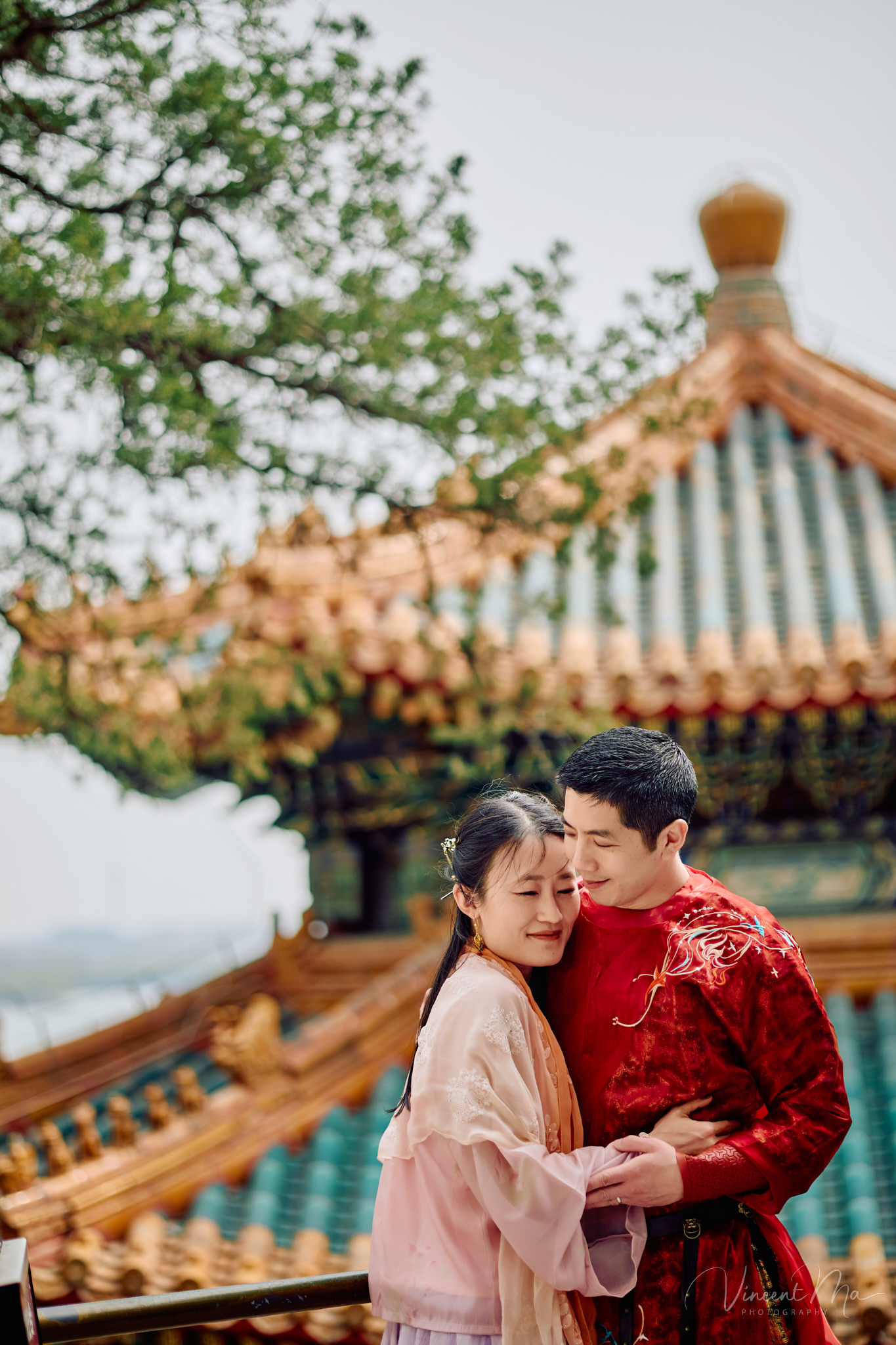 American couple in red and pink traditional Chinese clothing framed by delicate apricot blossoms during a Beijing spring photoshoot in the Summer palace.