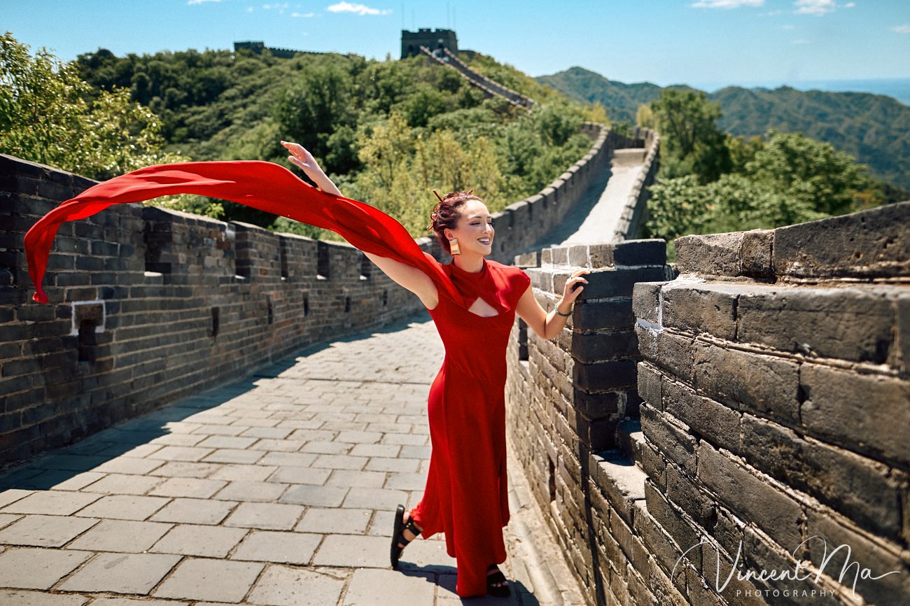Wide-angle cinematic fashion shot at the empty Mutianyu Great Wall. A solo traveler in a flowing dress silhouetted against the vast mountain ridges of Beijing.