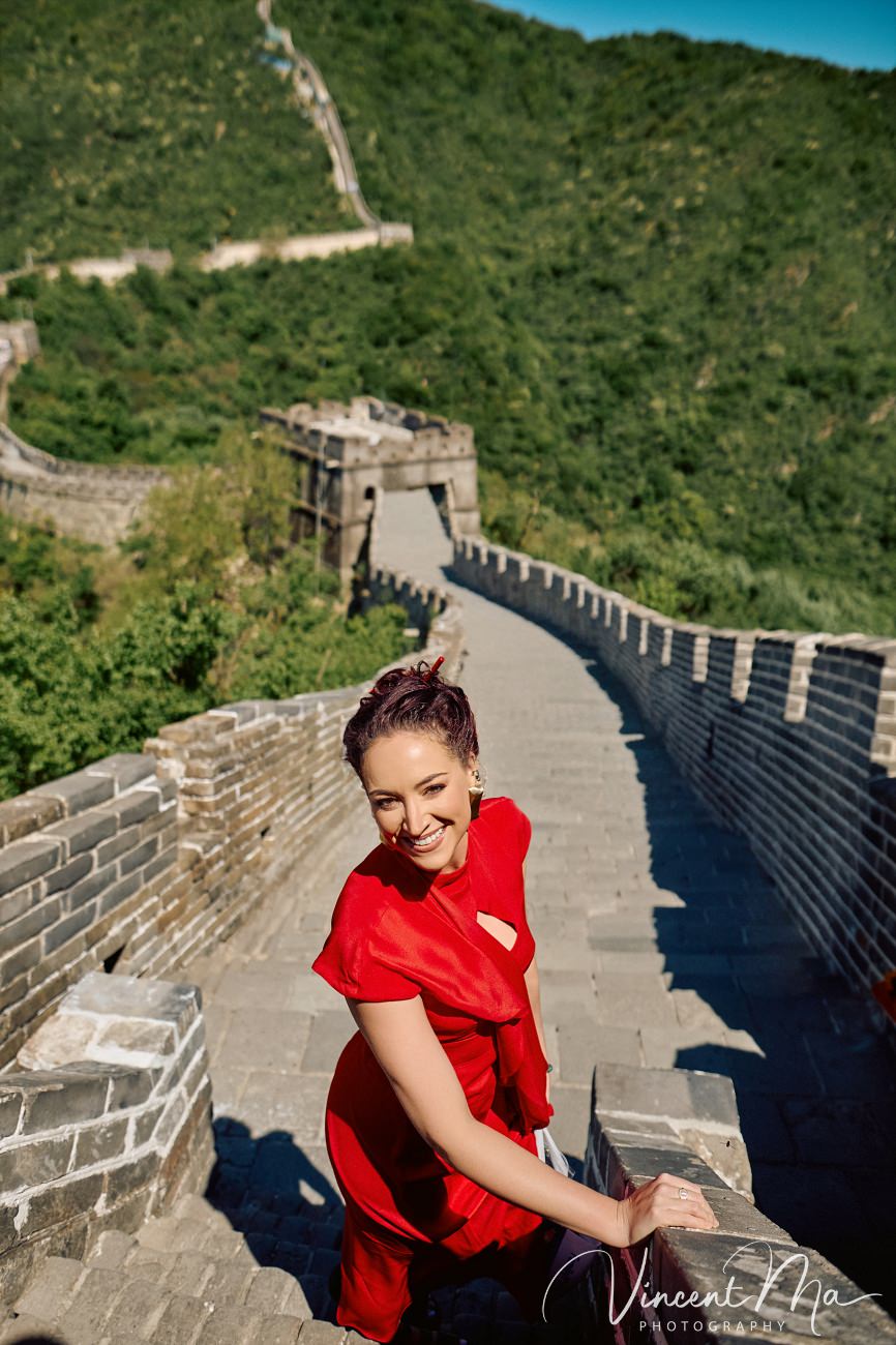 Wide-angle cinematic fashion shot at the empty Mutianyu Great Wall. A solo traveler in a flowing dress silhouetted against the vast mountain ridges of Beijing.