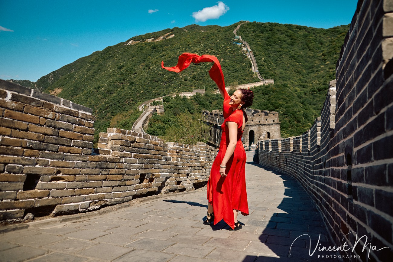 Wide-angle cinematic fashion shot at the empty Mutianyu Great Wall. A solo traveler in a flowing dress silhouetted against the vast mountain ridges of Beijing.