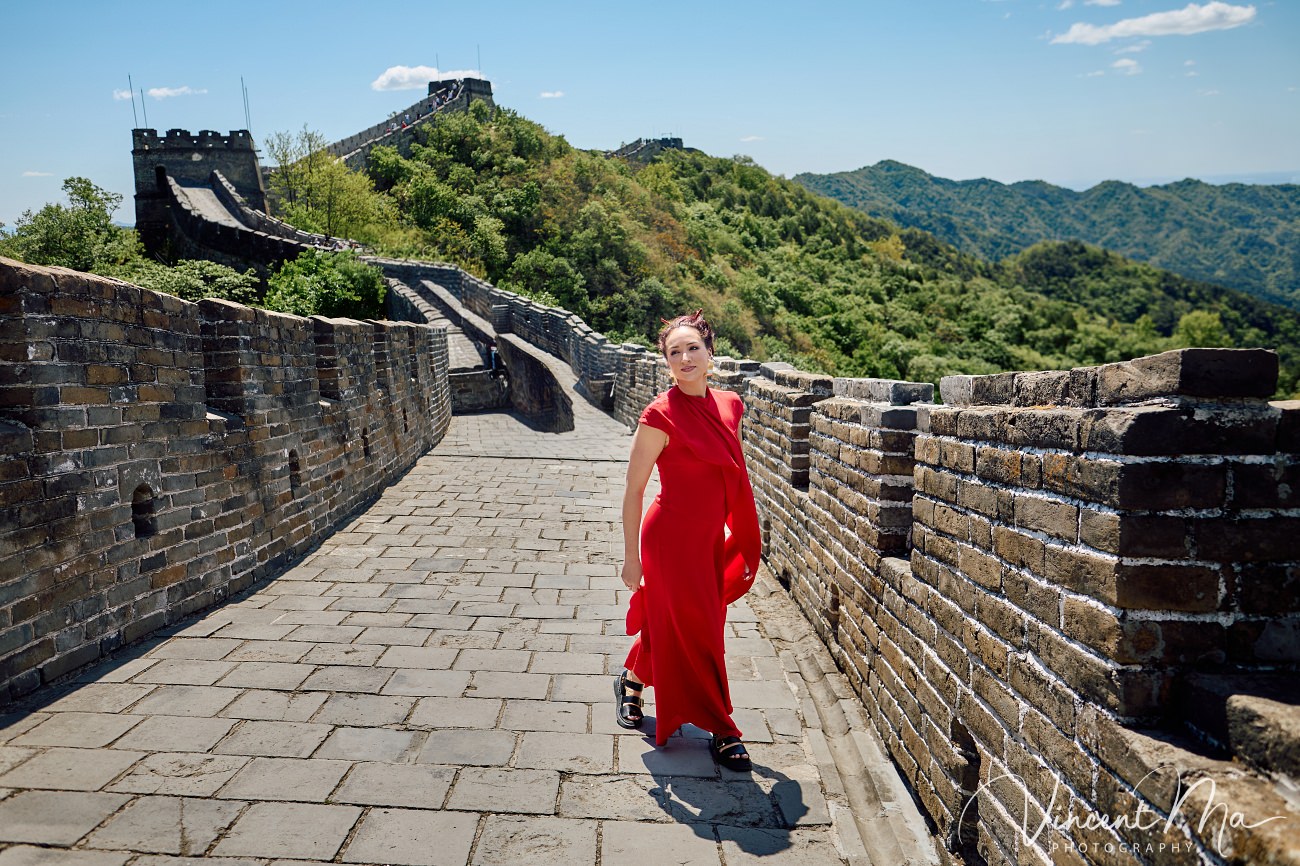 Wide-angle cinematic fashion shot at the empty Mutianyu Great Wall. A solo traveler in a flowing dress silhouetted against the vast mountain ridges of Beijing.