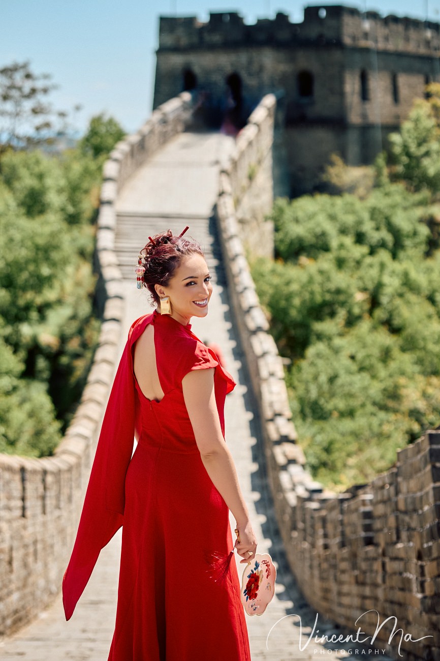 Wide-angle cinematic fashion shot at the empty Mutianyu Great Wall. A solo traveler in a flowing dress silhouetted against the vast mountain ridges of Beijing.