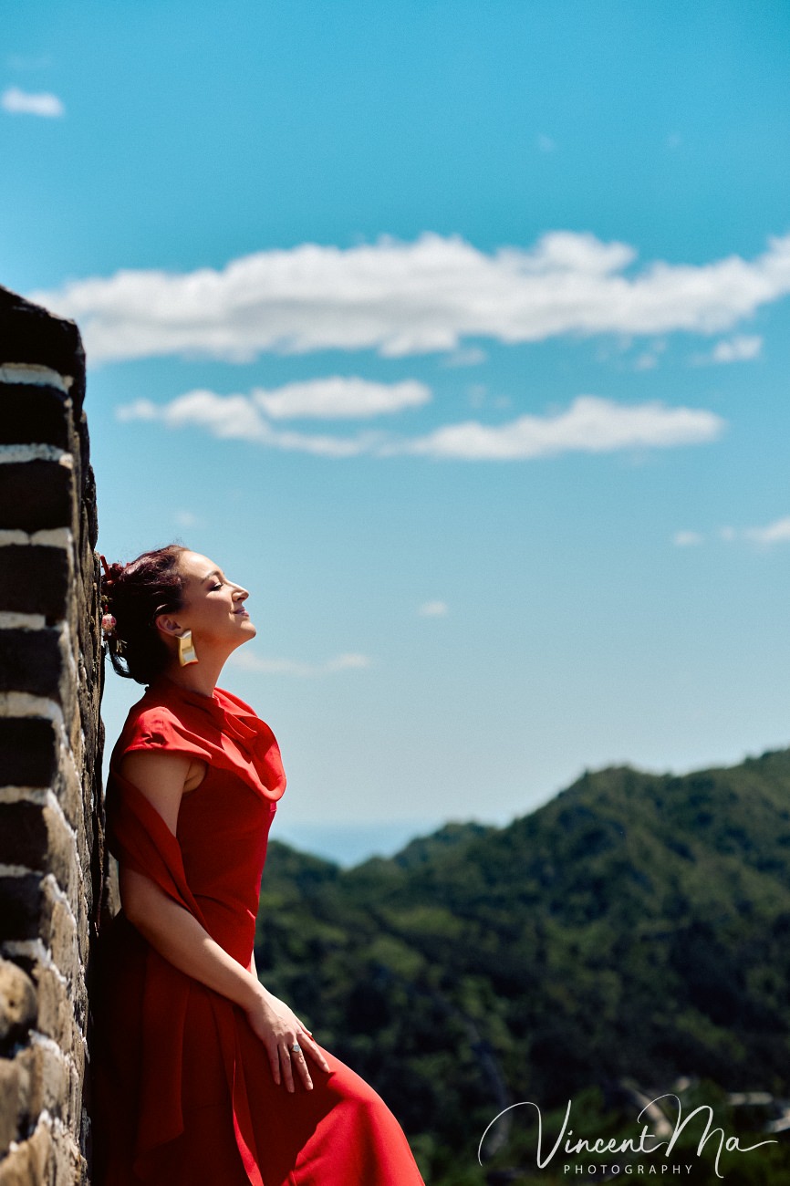 Wide-angle cinematic fashion shot at the empty Mutianyu Great Wall. A solo traveler in a flowing dress silhouetted against the vast mountain ridges of Beijing.