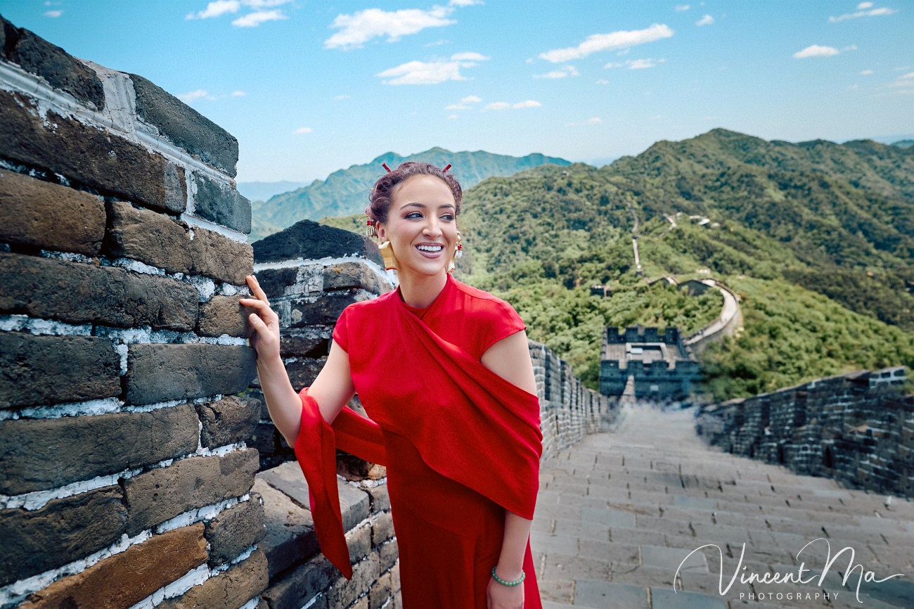 Wide-angle cinematic fashion shot at the empty Mutianyu Great Wall. A solo traveler in a flowing dress silhouetted against the vast mountain ridges of Beijing.