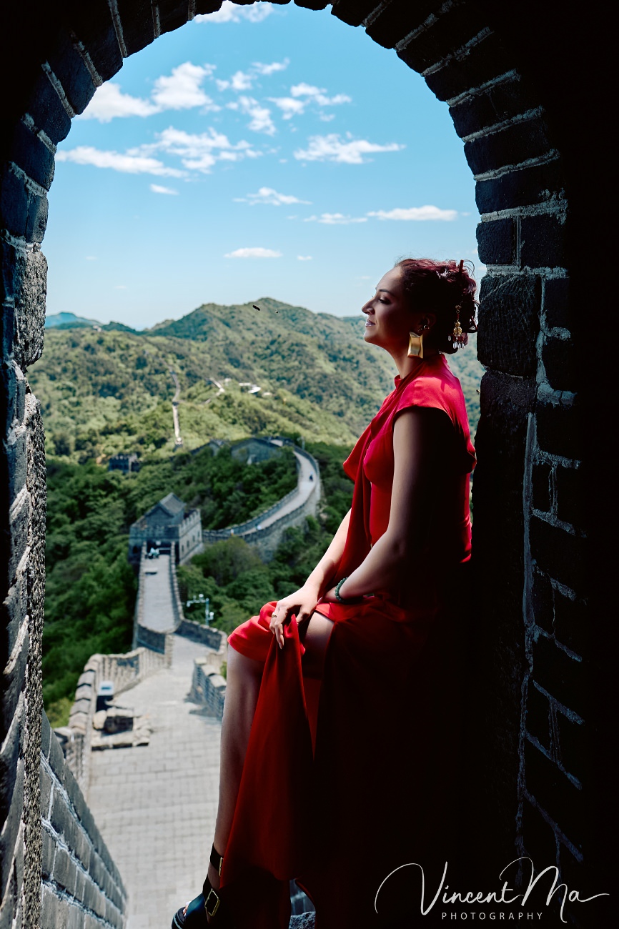 Wide-angle cinematic fashion shot at the empty Mutianyu Great Wall. A solo traveler in a flowing dress silhouetted against the vast mountain ridges of Beijing.