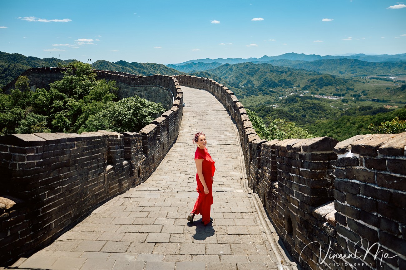 Wide-angle cinematic fashion shot at the empty Mutianyu Great Wall. A solo traveler in a flowing dress silhouetted against the vast mountain ridges of Beijing.
