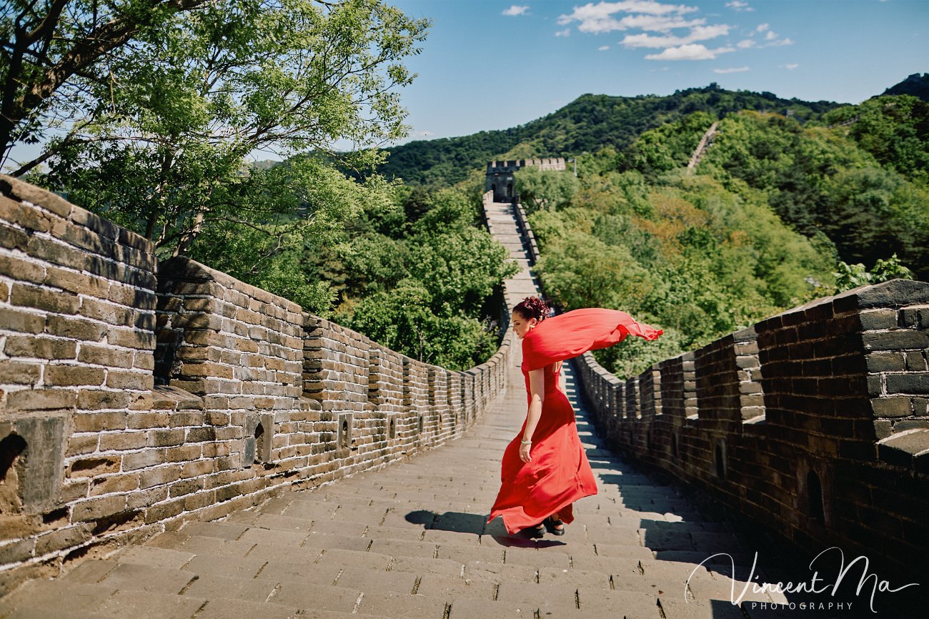 Wide-angle cinematic fashion shot at the empty Mutianyu Great Wall. A solo traveler in a flowing dress silhouetted against the vast mountain ridges of Beijing.